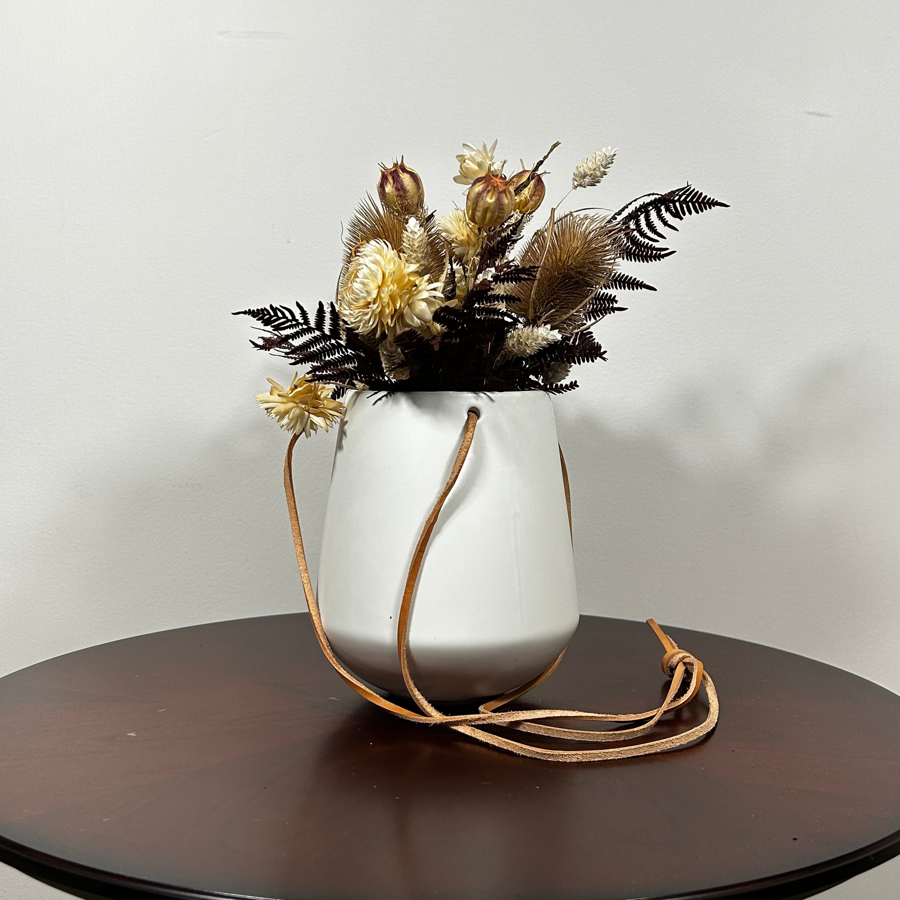 White hanging planter with beautiful dried flowers arrangement with burgundy bracken, ivory helichrysums, Nigel seed pods, natural teasels, and ivory phalaris on a beige background