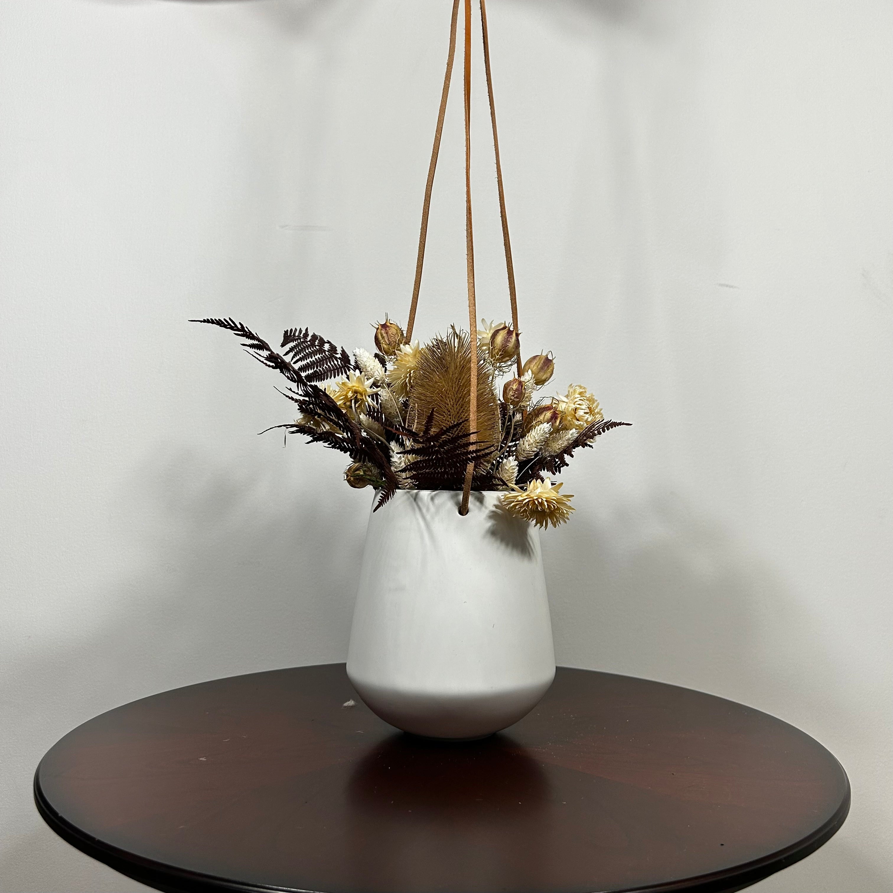 White hanging planter with beautiful dried flowers arrangement with burgundy bracken, ivory helichrysums, Nigel seed pods, natural teasels, and ivory phalaris on a beige background