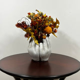 A dried flower arrangement in a concrete vessel sculpted to resemble four hands in a circle. The botanicals include preserved tones of yellow scarlet oak leaves and the deep, rich crimson of Chuisco Grass. Fiery Carthamus (safflower) and luminous Helichrysums (strawflowers) in golden and orange tones add a radiant glow. The composition is completed with a whimsical and charming detail: three small, natural reed grass pumpkins, perfectly nestled within the blooms.
