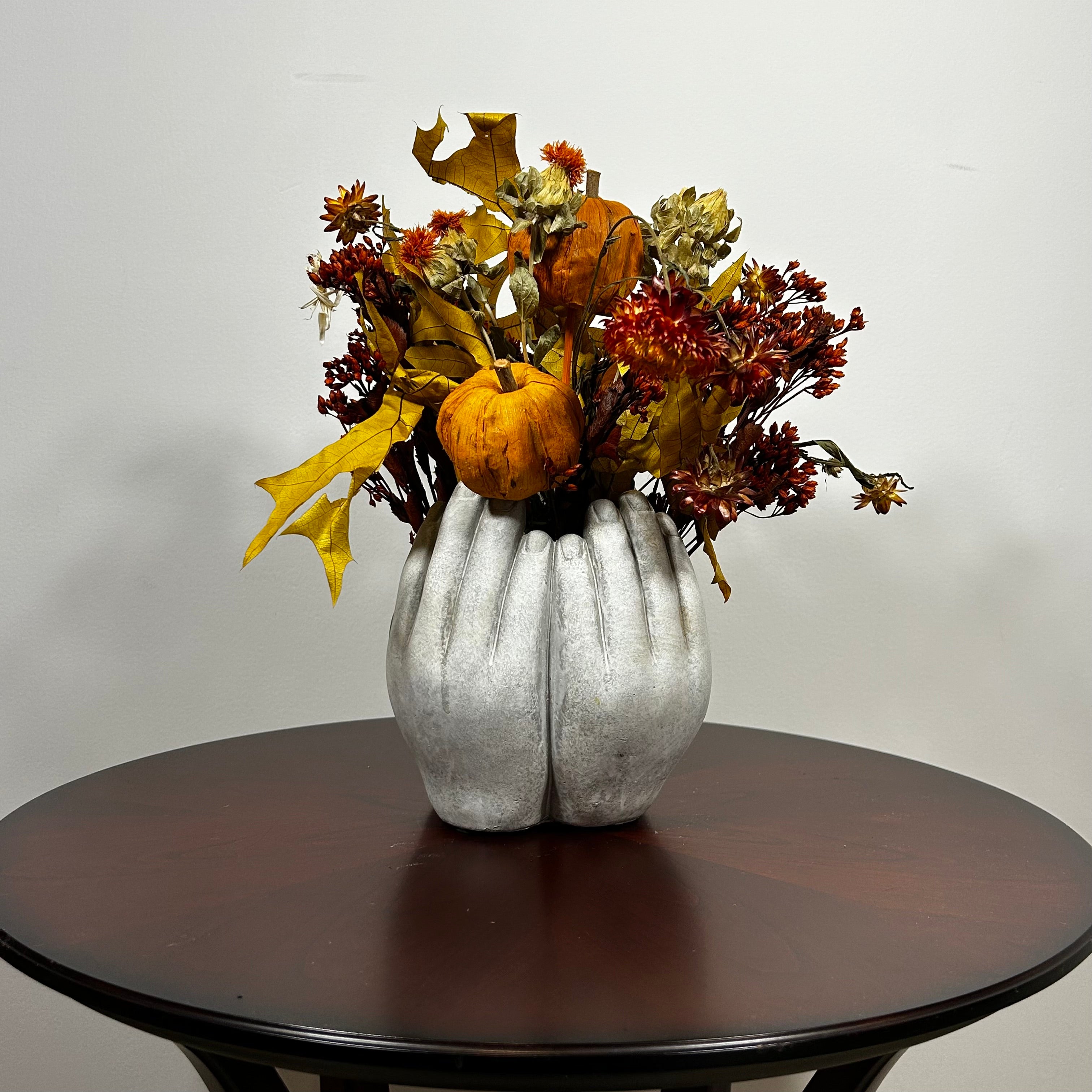A dried flower arrangement in a concrete vessel sculpted to resemble four hands in a circle. The botanicals include preserved tones of yellow scarlet oak leaves and the deep, rich crimson of Chuisco Grass. Fiery Carthamus (safflower) and luminous Helichrysums (strawflowers) in golden and orange tones add a radiant glow. The composition is completed with a whimsical and charming detail: three small, natural reed grass pumpkins, perfectly nestled within the blooms.