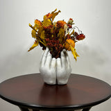 A dried flower arrangement in a concrete vessel sculpted to resemble four hands in a circle. The botanicals include preserved tones of yellow scarlet oak leaves and the deep, rich crimson of Chuisco Grass. Fiery Carthamus (safflower) and luminous Helichrysums (strawflowers) in golden and orange tones add a radiant glow. The composition is completed with a whimsical and charming detail: three small, natural reed grass pumpkins, perfectly nestled within the blooms.