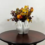 A dried flower arrangement in a concrete vessel sculpted to resemble four hands in a circle. The botanicals include preserved tones of yellow scarlet oak leaves and the deep, rich crimson of Chuisco Grass. Fiery Carthamus (safflower) and luminous Helichrysums (strawflowers) in golden and orange tones add a radiant glow. The composition is completed with a whimsical and charming detail: three small, natural reed grass pumpkins, perfectly nestled within the blooms.