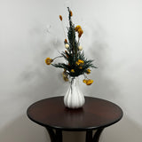 White modern ceramic vase filled with lush, deep green preserved Cedar. Rising from this foliage is a luminous cascade of golden Helichrysums (strawflowers) and vibrant yellow Achillea Parker (Yarrow), enhancing the arrangement's sun-kissed glow. These golden plumes are beautifully contrasted by the crisp, sculptural spheres of bleached white Craspedia and playful white curly ting ting. The arrangement is on a dark wooden table against a light grey background