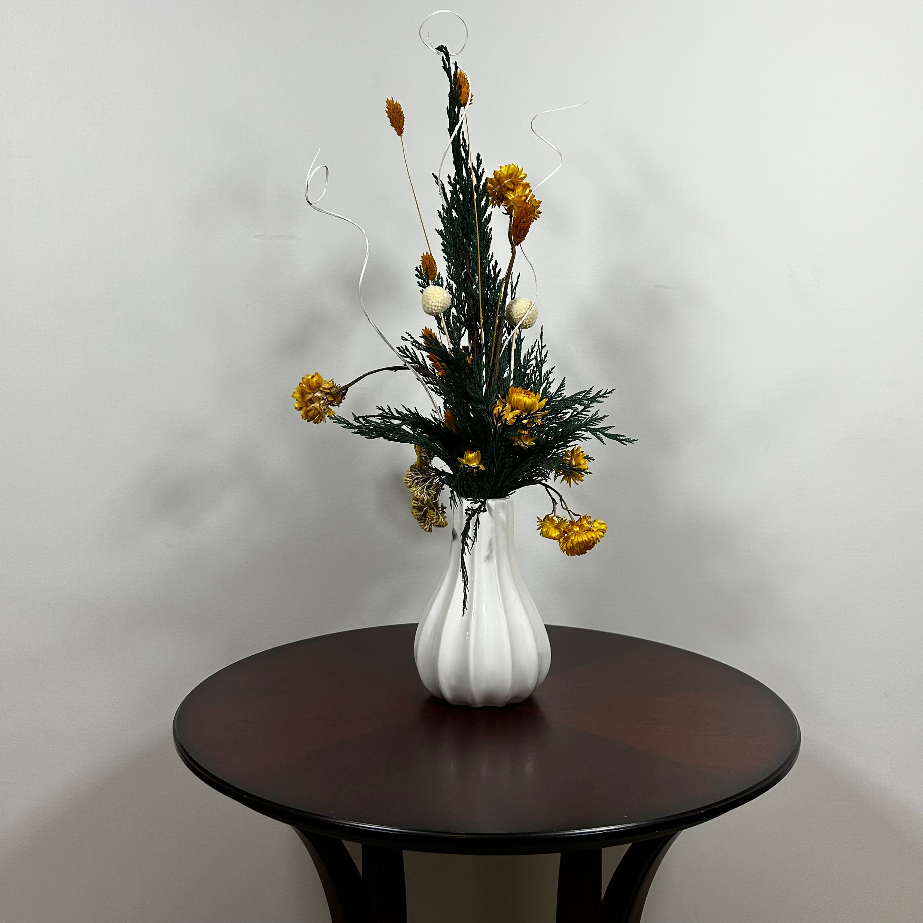 White modern ceramic vase filled with lush, deep green preserved Cedar. Rising from this foliage is a luminous cascade of golden Helichrysums (strawflowers) and vibrant yellow Achillea Parker (Yarrow), enhancing the arrangement's sun-kissed glow. These golden plumes are beautifully contrasted by the crisp, sculptural spheres of bleached white Craspedia and playful white curly ting ting. The arrangement is on a dark wooden table against a light grey background