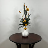 White modern ceramic vase filled with lush, deep green preserved Cedar. Rising from this foliage is a luminous cascade of golden Helichrysums (strawflowers) and vibrant yellow Achillea Parker (Yarrow), enhancing the arrangement's sun-kissed glow. These golden plumes are beautifully contrasted by the crisp, sculptural spheres of bleached white Craspedia and playful white curly ting ting. The arrangement is on a dark wooden table against a light grey background