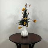 White modern ceramic vase filled with lush, deep green preserved Cedar. Rising from this foliage is a luminous cascade of golden Helichrysums (strawflowers) and vibrant yellow Achillea Parker (Yarrow), enhancing the arrangement's sun-kissed glow. These golden plumes are beautifully contrasted by the crisp, sculptural spheres of bleached white Craspedia and playful white curly ting ting. The arrangement is on a dark wooden table against a light grey background