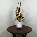 White modern ceramic vase filled with lush, deep green preserved Cedar. Rising from this foliage is a luminous cascade of golden Helichrysums (strawflowers) and vibrant yellow Achillea Parker (Yarrow), enhancing the arrangement's sun-kissed glow. These golden plumes are beautifully contrasted by the crisp, sculptural spheres of bleached white Craspedia and playful white curly ting ting. The arrangement is on a dark wooden table against a light grey background