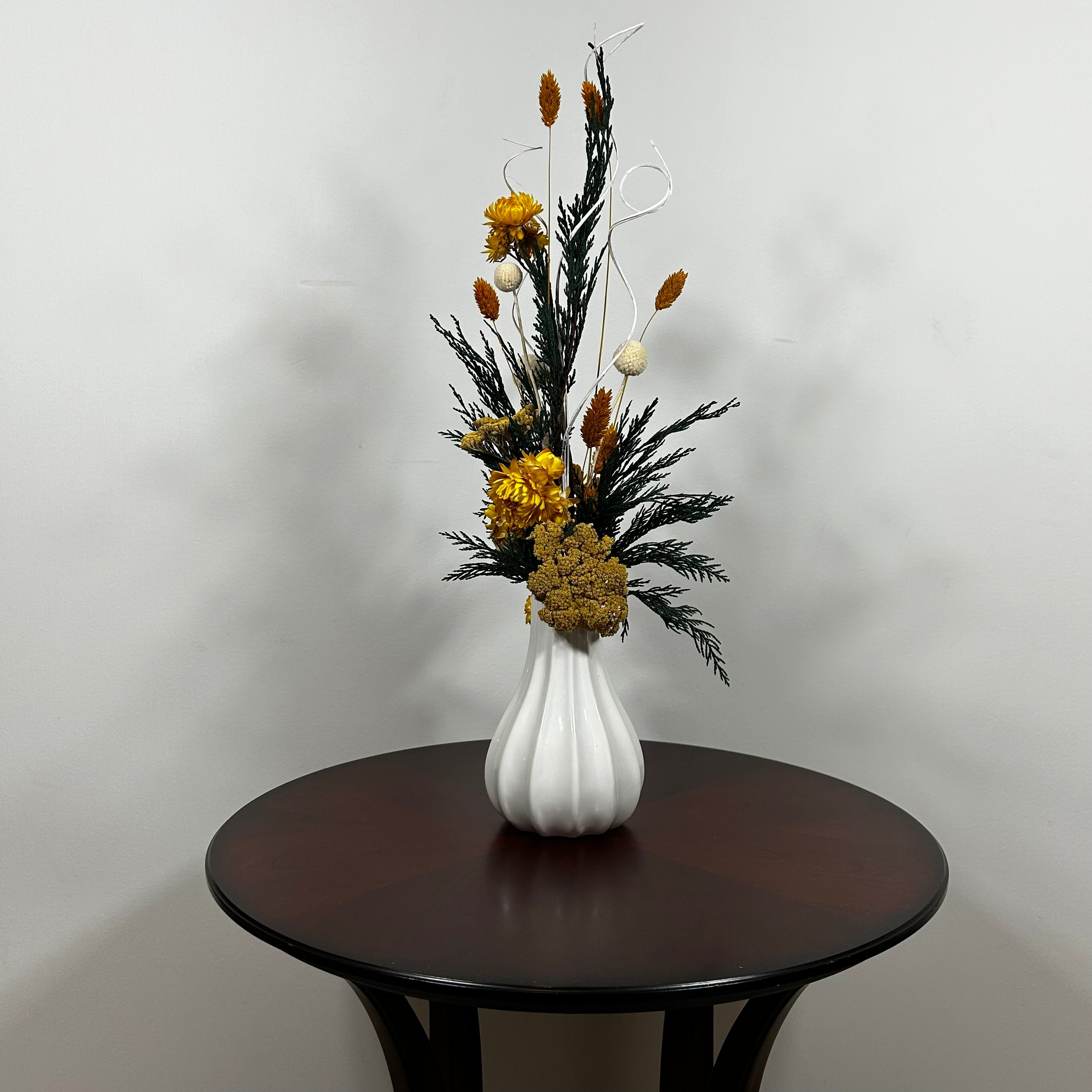 White modern ceramic vase filled with lush, deep green preserved Cedar. Rising from this foliage is a luminous cascade of golden Helichrysums (strawflowers) and vibrant yellow Achillea Parker (Yarrow), enhancing the arrangement's sun-kissed glow. These golden plumes are beautifully contrasted by the crisp, sculptural spheres of bleached white Craspedia and playful white curly ting ting. The arrangement is on a dark wooden table against a light grey background