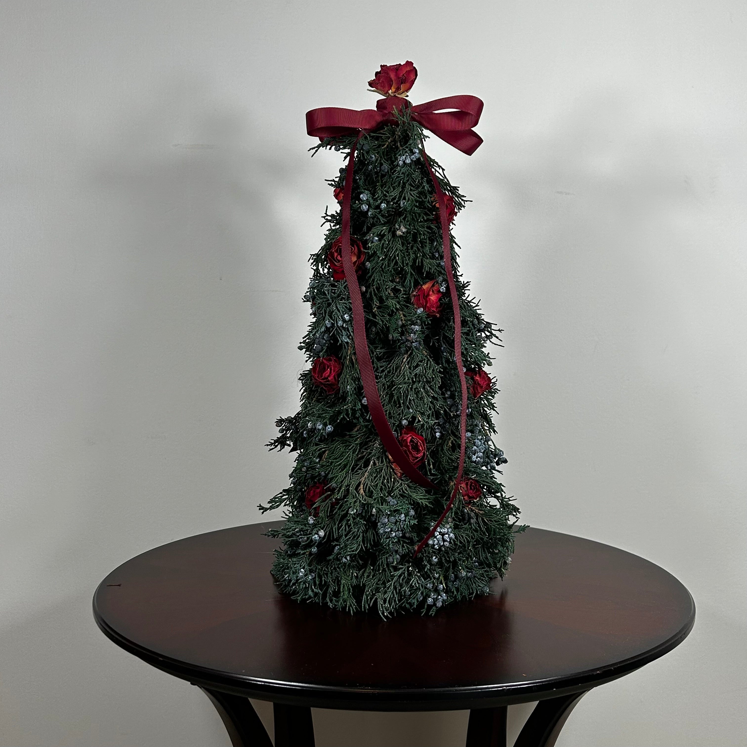 Decorative Christmas tree this 18-inch design is densely layered with fragrant, preserved Juniper and its signature blue berries, with beautiful deep red "Bright Torch" roses are placed to command attention. The entire piece is finished with a large, elegant deep red grosgrain double bow, whose ribbons cascade down.
