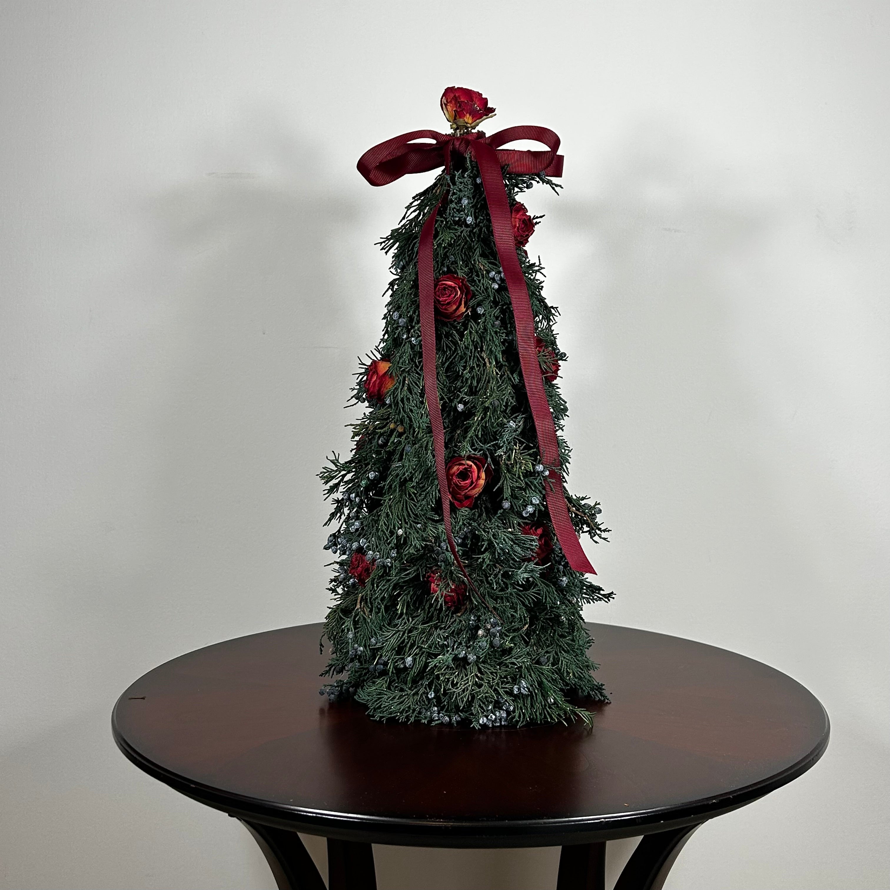 Decorative Christmas tree this 18-inch design is densely layered with fragrant, preserved Juniper and its signature blue berries, with beautiful deep red "Bright Torch" roses are placed to command attention. The entire piece is finished with a large, elegant deep red grosgrain double bow, whose ribbons cascade down.