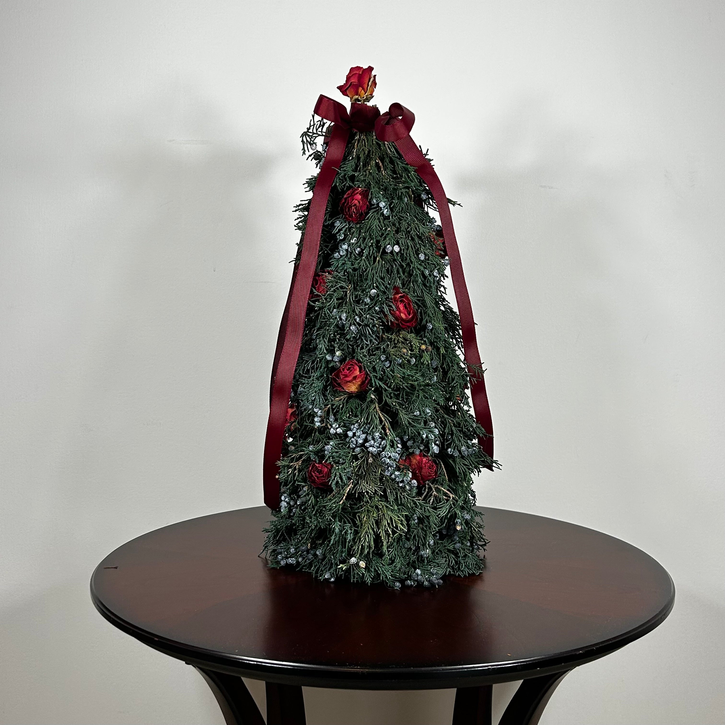 Decorative Christmas tree this 18-inch design is densely layered with fragrant, preserved Juniper and its signature blue berries, with beautiful deep red "Bright Torch" roses are placed to command attention. The entire piece is finished with a large, elegant deep red grosgrain double bow, whose ribbons cascade down.