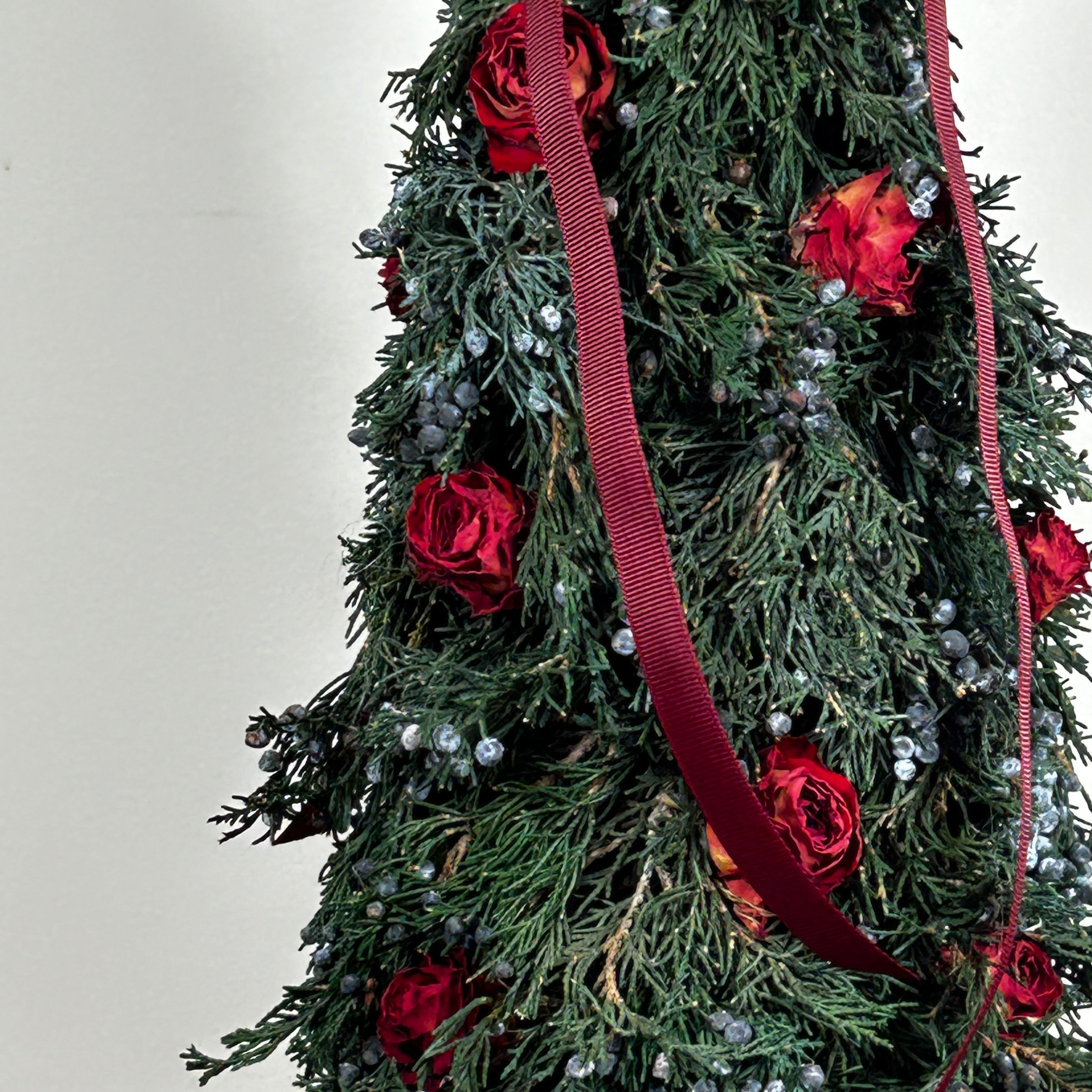 Close-up of a decorative Christmas tree this 18-inch design is densely layered with fragrant, preserved Juniper and its signature blue berries, with beautiful deep red "Bright Torch" roses are placed to command attention. The entire piece is finished with a large, elegant deep red grosgrain double bow, whose ribbons cascade down.