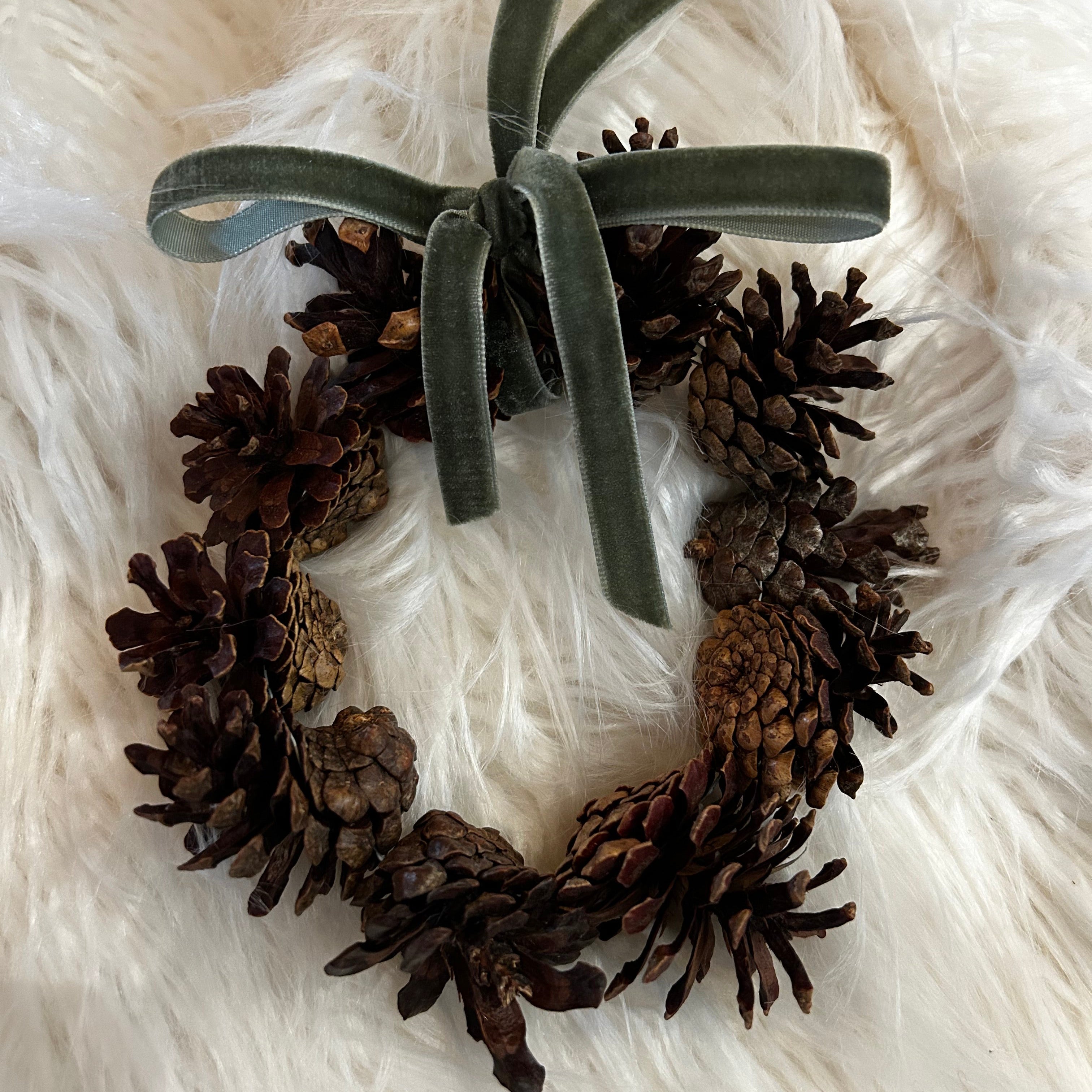 A miniature wreath in the rustic tones of the pinecones, that are beautifully complemented by a delicate, soft green ribbon tied into a small, elegant bow that doubles as the hanging loop.