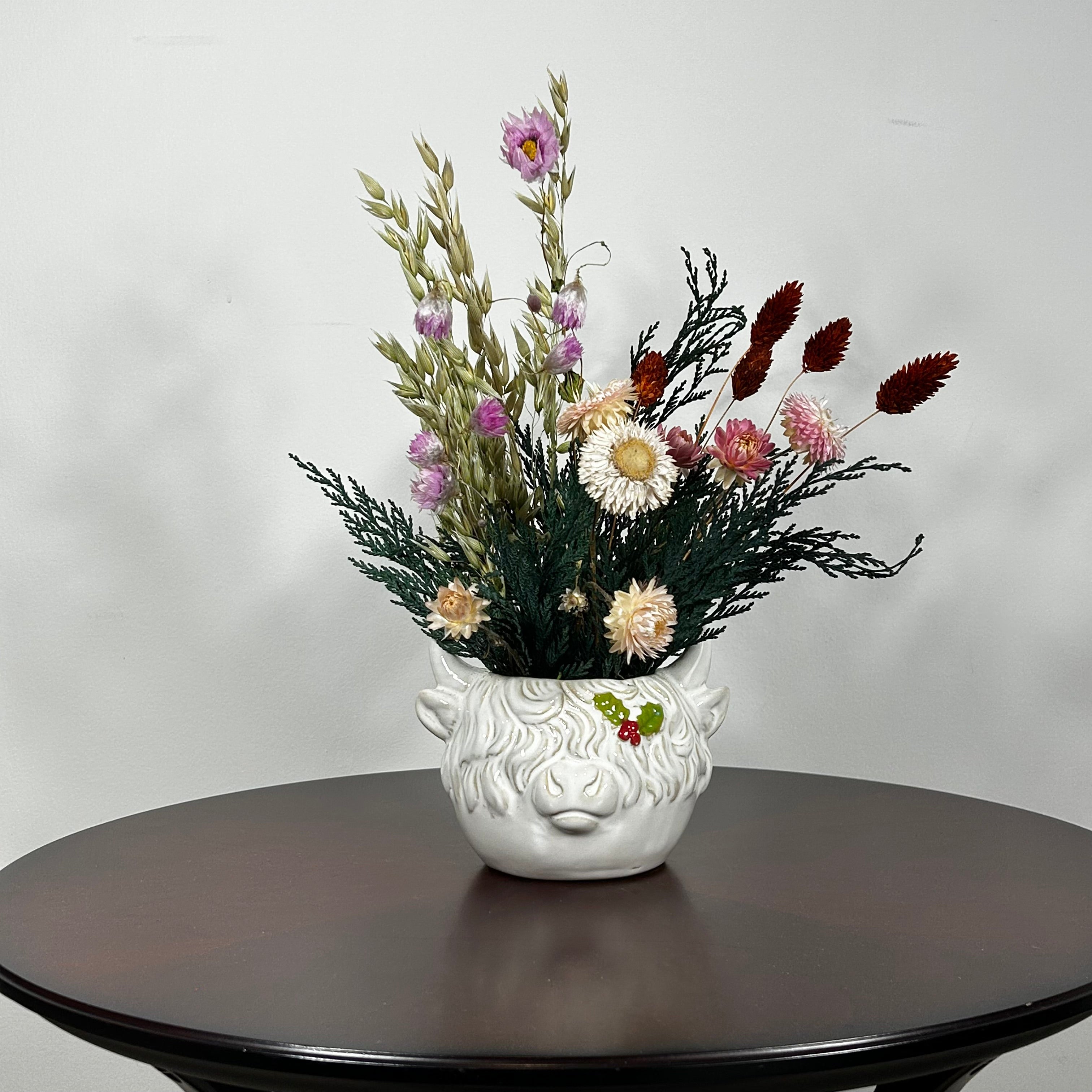 A matt white ceramic planter in the shape of a Highland Cow's head holds a rustic arrangement that features the rich green texture of preserved Cedar, providing a classic holiday base. This is layered with the soft textures of natural Quaker Oats, the gentle colour of natural pink Helichrysums, and the vivid contrast of burgundy dyed Phalaris. Floating delicately above the bulk of the arrangement are ethereal paper daisies in natural pink, adding a final touch of whimsy.