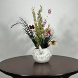 A matt white ceramic planter in the shape of a Highland Cow's head holds a rustic arrangement that features the rich green texture of preserved Cedar, providing a classic holiday base. This is layered with the soft textures of natural Quaker Oats, the gentle colour of natural pink Helichrysums, and the vivid contrast of burgundy dyed Phalaris. Floating delicately above the bulk of the arrangement are ethereal paper daisies in natural pink, adding a final touch of whimsy.