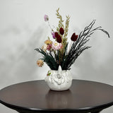 A matt white ceramic planter in the shape of a Highland Cow's head holds a rustic arrangement that features the rich green texture of preserved Cedar, providing a classic holiday base. This is layered with the soft textures of natural Quaker Oats, the gentle colour of natural pink Helichrysums, and the vivid contrast of burgundy dyed Phalaris. Floating delicately above the bulk of the arrangement are ethereal paper daisies in natural pink, adding a final touch of whimsy.