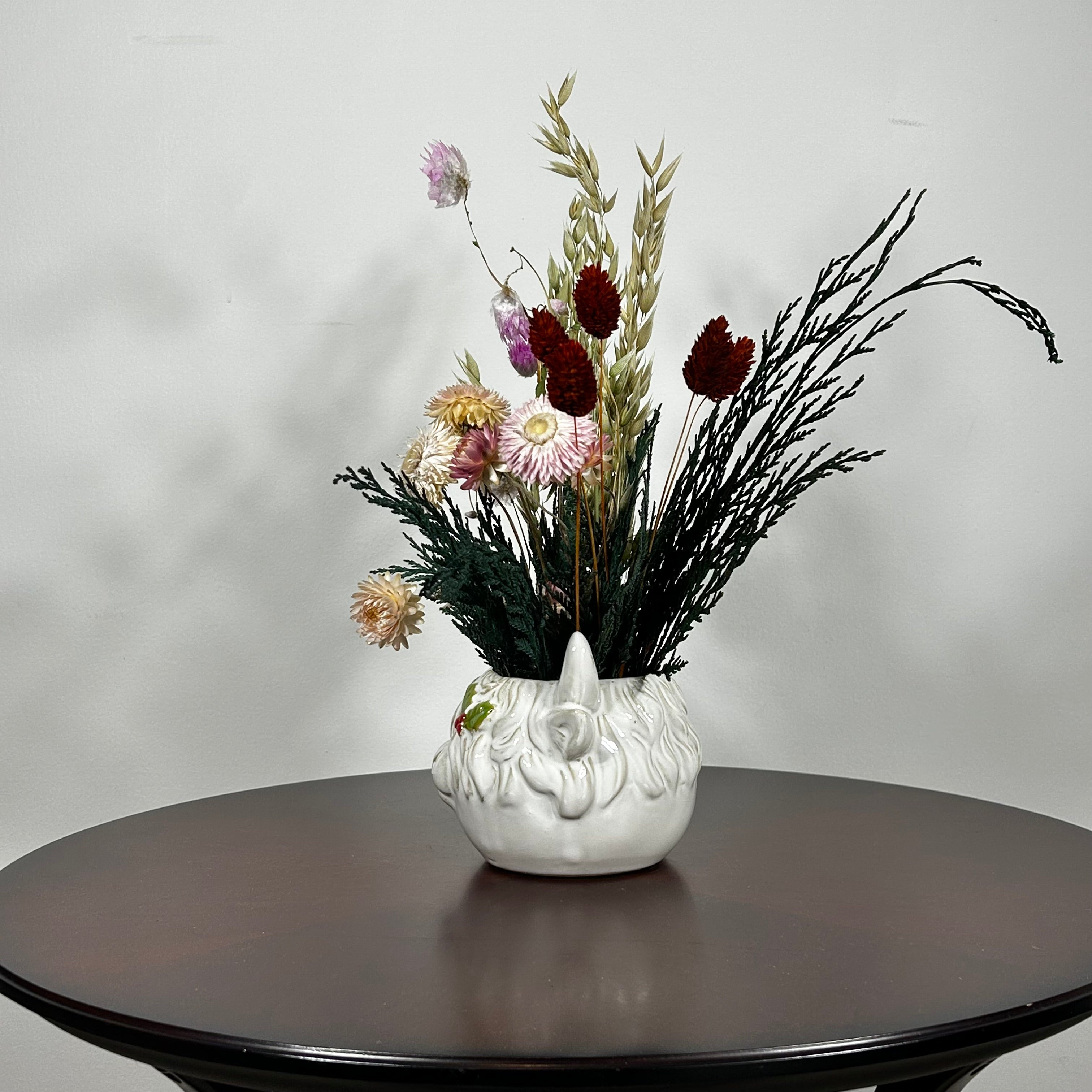 A matt white ceramic planter in the shape of a Highland Cow's head holds a rustic arrangement that features the rich green texture of preserved Cedar, providing a classic holiday base. This is layered with the soft textures of natural Quaker Oats, the gentle colour of natural pink Helichrysums, and the vivid contrast of burgundy dyed Phalaris. Floating delicately above the bulk of the arrangement are ethereal paper daisies in natural pink, adding a final touch of whimsy.