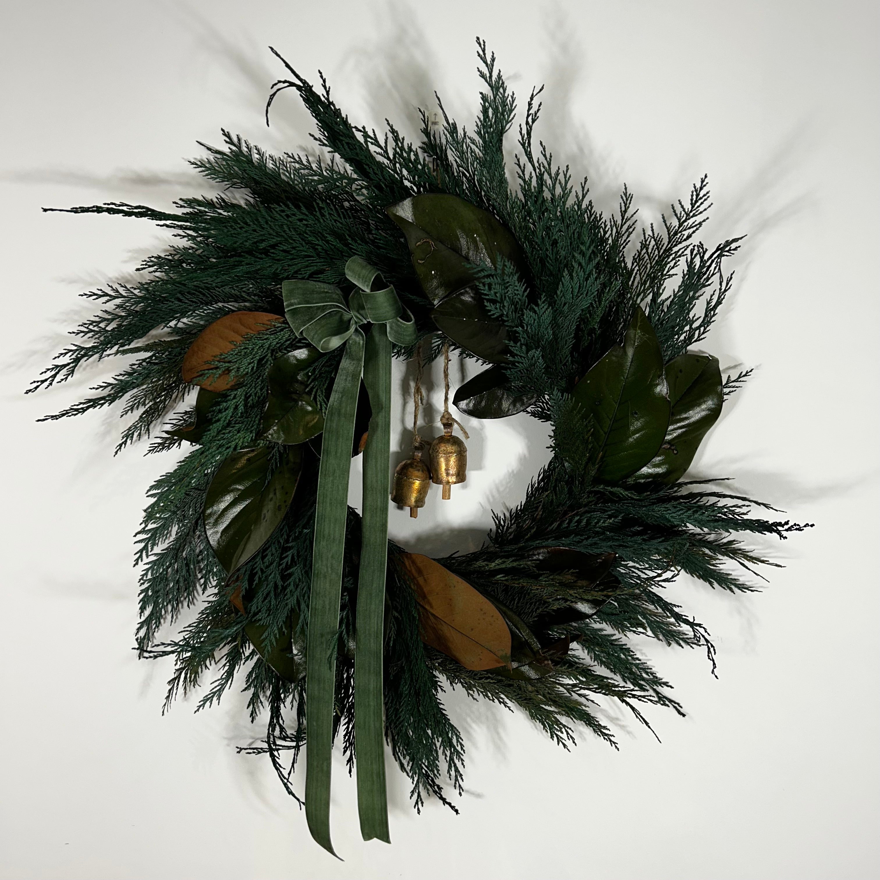 An Everlasting Wreath with striking contrast between the traditional, needle-like texture of preserved Cedar and the large, smooth, often bi-coloured leaves of preserved Magnolia. A soft green velvet bow is thoughtfully tied in the top left corner, adding a subtle touch of richness and colour. The design is perfected by the gentle chime of two small vintage iron bells, which hang suspended in the centre by natural jute rope.