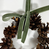 A close-up view of a miniature wreath in the rustic tones of the pinecones, that are beautifully complemented by a delicate, soft green ribbon tied into a small, elegant bow that doubles as the hanging loop.