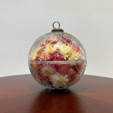 A striking 6-inch ornament is crafted from clear glass, featuring a detailed etched diamond pattern with flower shapes nestled inside, lending it a unique, vintage charm. The sphere is capped with a traditional ornament hanger, finished in antiqued bronze, lending to its precious, heirloom look. Inside, the sphere is filled with beautiful natural pink Helichrysums (strawflowers), whose delicate colors are softly magnified by the glass.