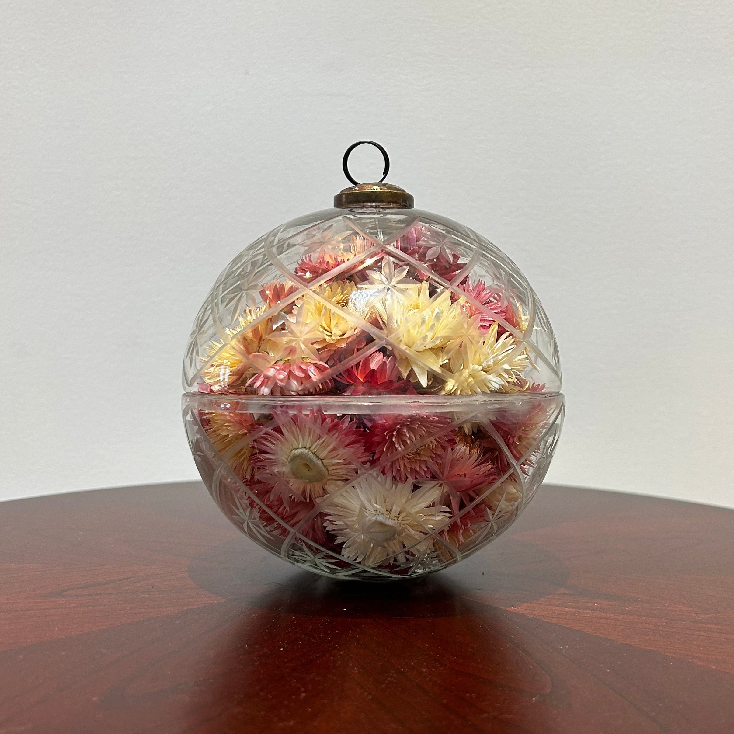 A striking 6-inch ornament is crafted from clear glass, featuring a detailed etched diamond pattern with flower shapes nestled inside, lending it a unique, vintage charm. The sphere is capped with a traditional ornament hanger, finished in antiqued bronze, lending to its precious, heirloom look. Inside, the sphere is filled with beautiful natural pink Helichrysums (strawflowers), whose delicate colors are softly magnified by the glass.