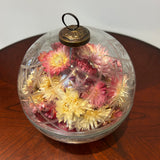 A striking 6-inch ornament is crafted from clear glass, featuring a detailed etched diamond pattern with flower shapes nestled inside, lending it a unique, vintage charm. The sphere is capped with a traditional ornament hanger, finished in antiqued bronze, lending to its precious, heirloom look. Inside, the sphere is filled with beautiful natural pink Helichrysums (strawflowers), whose delicate colors are softly magnified by the glass.