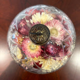 A striking 6-inch ornament is crafted from clear glass, featuring a detailed etched diamond pattern with flower shapes nestled inside, lending it a unique, vintage charm. The sphere is capped with a traditional ornament hanger, finished in antiqued bronze, lending to its precious, heirloom look. Inside, the sphere is filled with beautiful natural pink Helichrysums (strawflowers), whose delicate colors are softly magnified by the glass.