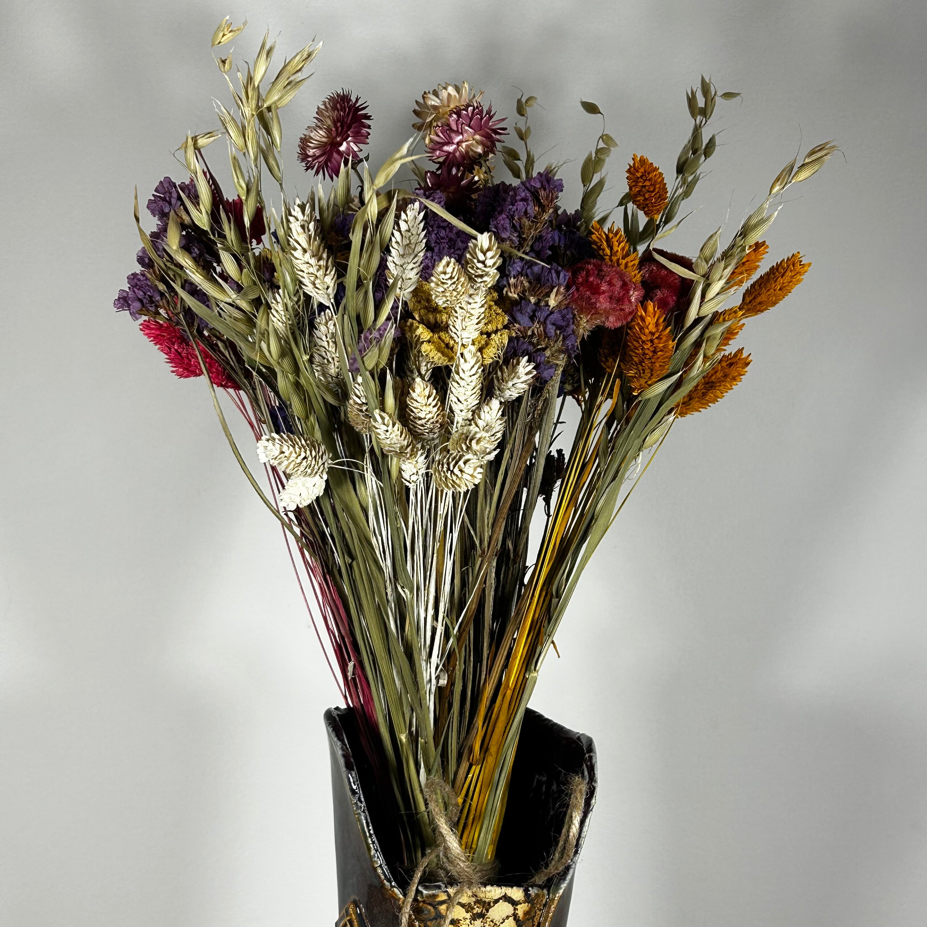 This magnificent, hand-tied bouquet is a vibrant symphony of contrasting colours and textures designed for the sophisticated interior. The Amethyst base of lush purple Statice provides a dramatic backdrop for the Gold accents: bright yellow Achillea Parker and soft natural pink Helichrysums. Textural elements featuring the unique, wavy shapes of red coral Celosia and Phalaris dyed in three complementary colors (natural, orange, and red).