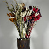 This magnificent, hand-tied bouquet is a vibrant symphony of contrasting colours and textures designed for the sophisticated interior. The Amethyst base of lush purple Statice provides a dramatic backdrop for the Gold accents: bright yellow Achillea Parker and soft natural pink Helichrysums. Textural elements featuring the unique, wavy shapes of red coral Celosia and Phalaris dyed in three complementary colors (natural, orange, and red).