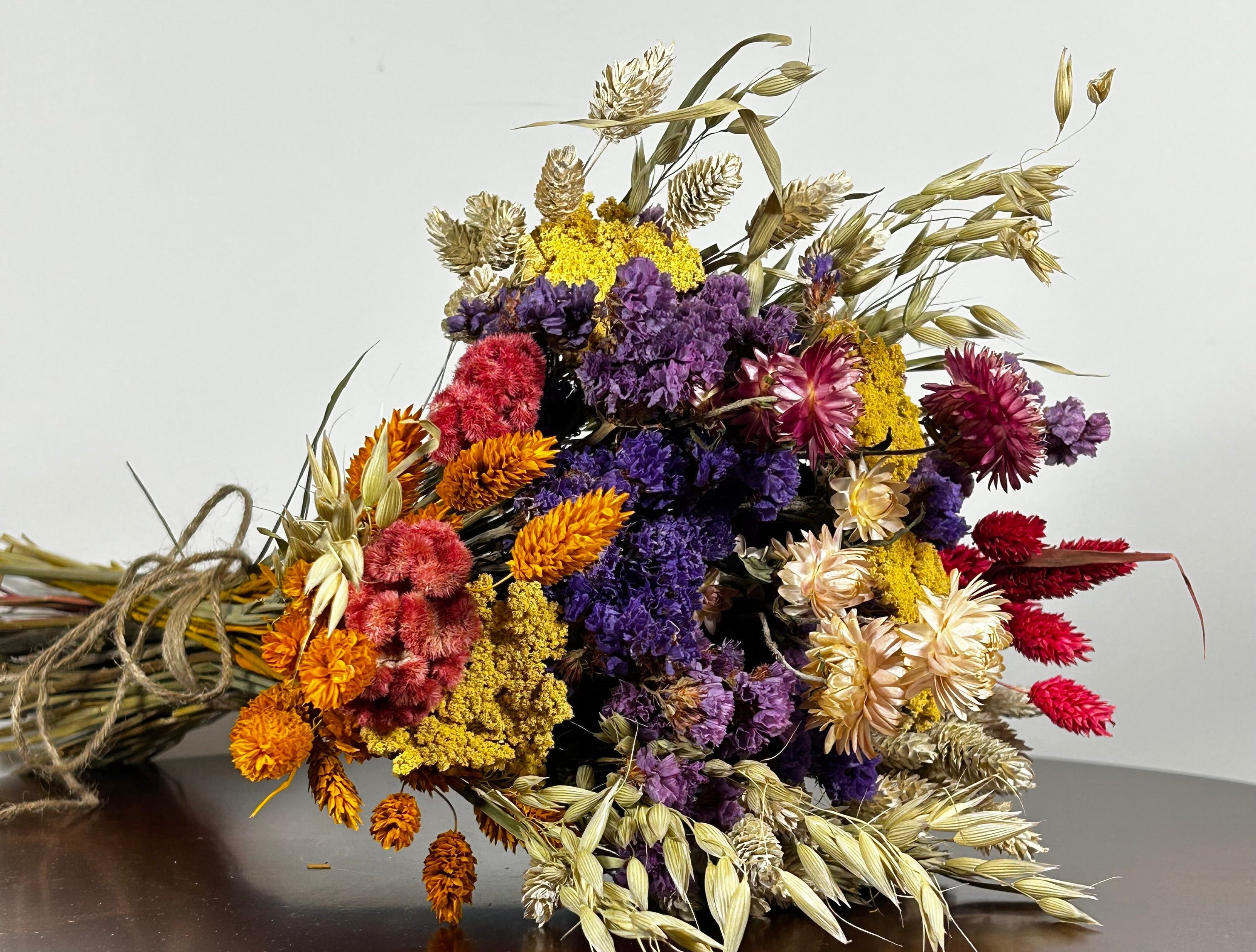 This magnificent, hand-tied bouquet is a vibrant symphony of contrasting colours and textures designed for the sophisticated interior. The Amethyst base of lush purple Statice provides a dramatic backdrop for the Gold accents: bright yellow Achillea Parker and soft natural pink Helichrysums. Textural elements featuring the unique, wavy shapes of red coral Celosia and Phalaris dyed in three complementary colors (natural, orange, and red).