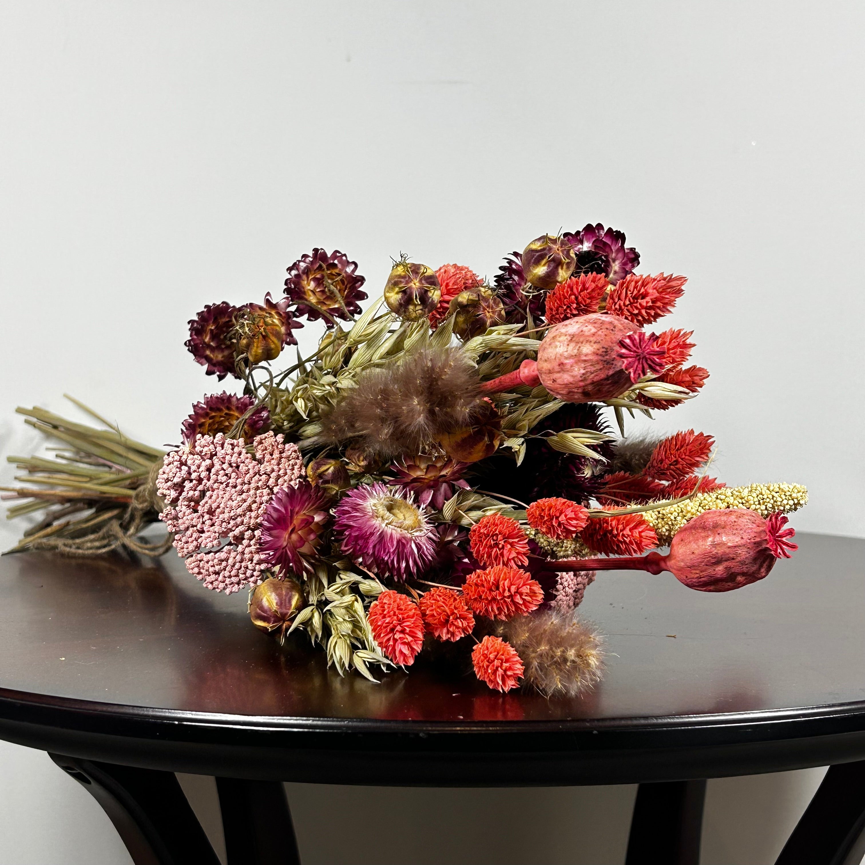 A hand-tied bouquet features the majestic, airy softness of natural Pampas Grass, contrasted with the deep, plush tones of deep burgundy Helichrysums.
The palette is enhanced by soft natural pink Helichrysums and vibrant dyed pink Achillea Parker, while coral-dyed Phalaris adds vertical movement. The rustic charm of burgundy Nigella seed pods, textured Poppy seed pods, and Quaker Oats provides intricate detail.