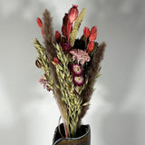 A hand-tied bouquet features the majestic, airy softness of natural Pampas Grass, contrasted with the deep, plush tones of deep burgundy Helichrysums.
The palette is enhanced by soft natural pink Helichrysums and vibrant dyed pink Achillea Parker, while coral-dyed Phalaris adds vertical movement. The rustic charm of burgundy Nigella seed pods, textured Poppy seed pods, and Quaker Oats provides intricate detail.