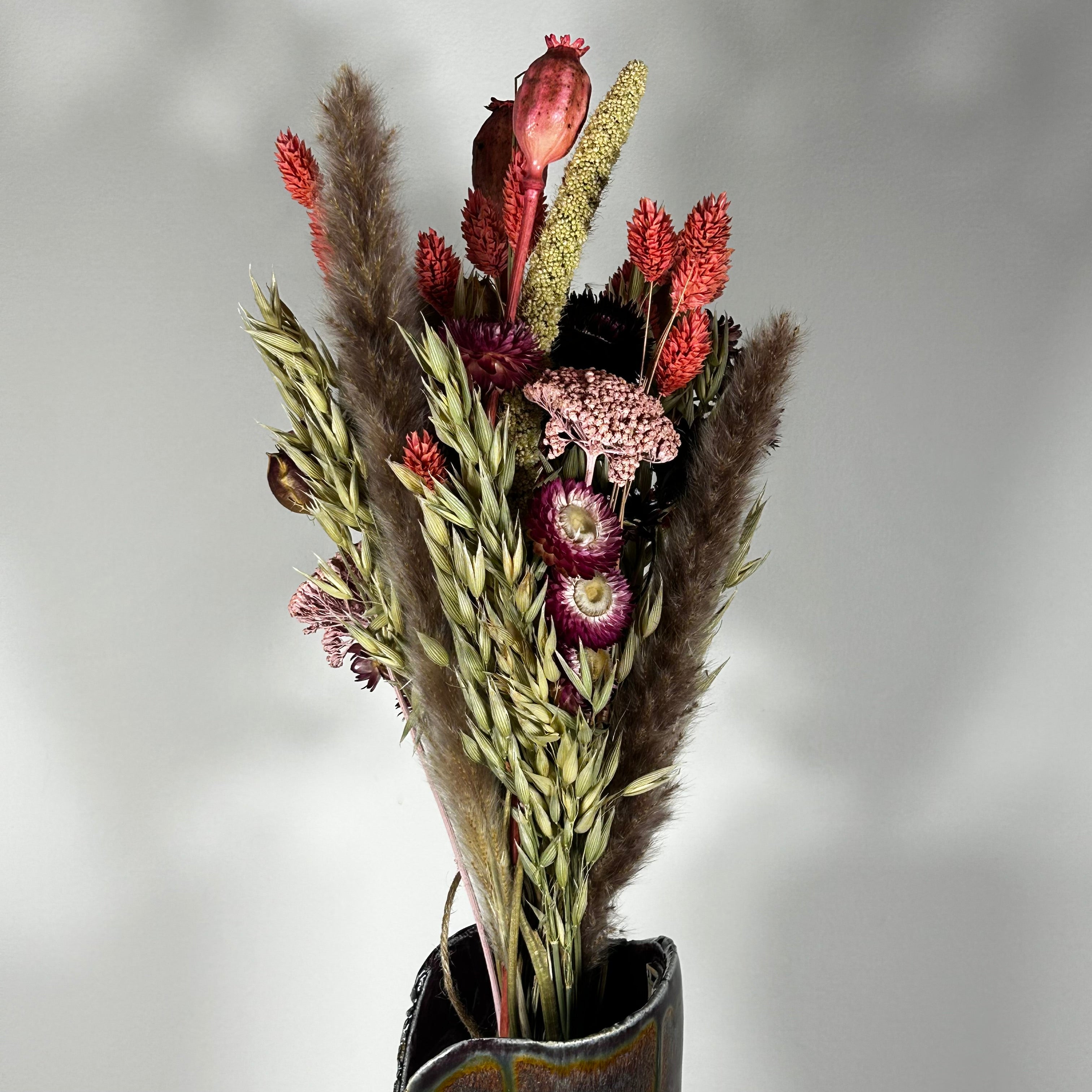 A hand-tied bouquet features the majestic, airy softness of natural Pampas Grass, contrasted with the deep, plush tones of deep burgundy Helichrysums.
The palette is enhanced by soft natural pink Helichrysums and vibrant dyed pink Achillea Parker, while coral-dyed Phalaris adds vertical movement. The rustic charm of burgundy Nigella seed pods, textured Poppy seed pods, and Quaker Oats provides intricate detail.