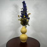 A yellow ribbed ceramic vase with a double-gourd shape, filled with artificial blue and yellow flowers, displayed on a dark brown wooden table against a plain white wall.
