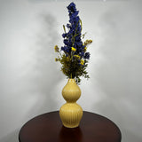 A yellow ribbed ceramic vase with a double-gourd shape, filled with artificial blue and yellow flowers, displayed on a dark brown wooden table against a plain white wall.