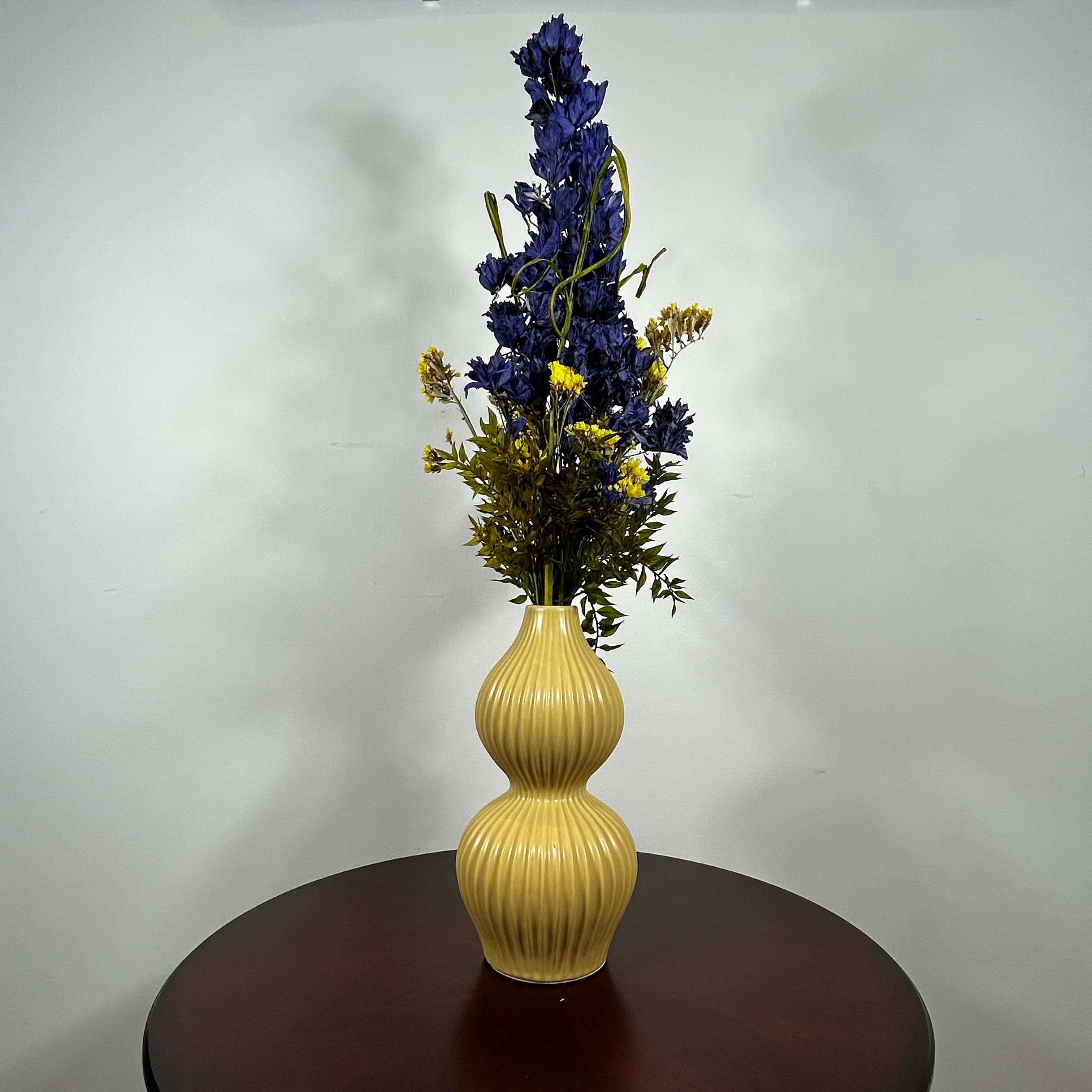 A yellow ribbed ceramic vase with a double-gourd shape, filled with artificial blue and yellow flowers, displayed on a dark brown wooden table against a plain white wall.