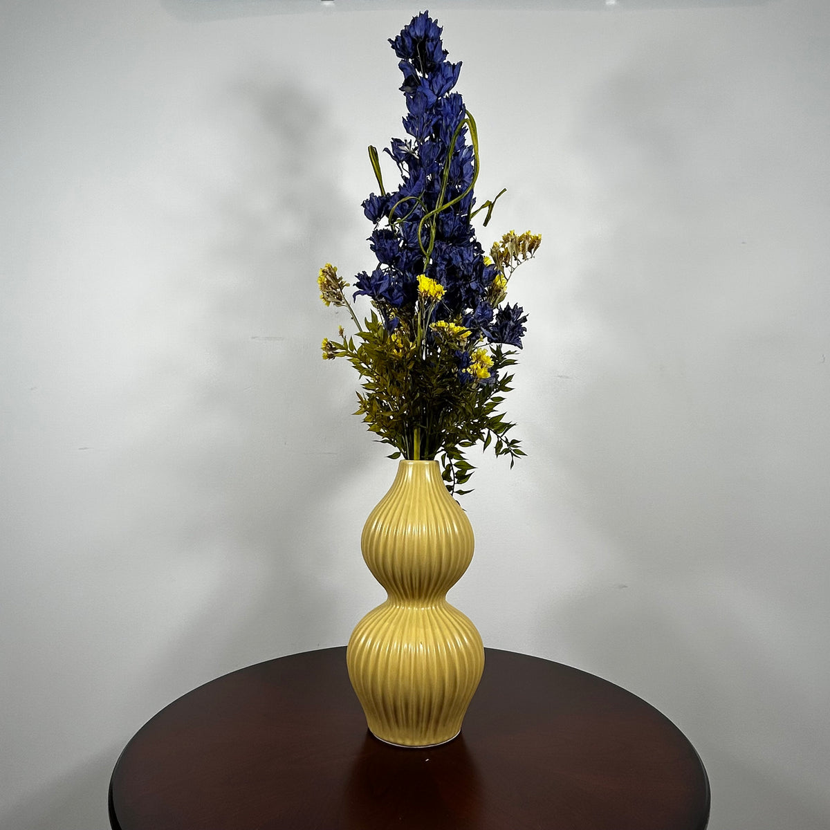 A yellow ribbed ceramic vase with a double-gourd shape, filled with artificial blue and yellow flowers, displayed on a dark brown wooden table against a plain white wall.