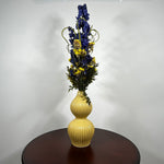 A dried floral arrangement featuring blue delphiniums, yellow flowers, and green foliage in a ribbed, two-bulb yellow ceramic vase, displayed on a dark wooden table against a plain white wall.