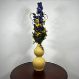 A dried floral arrangement featuring blue delphiniums, yellow flowers, and green foliage in a ribbed, two-bulb yellow ceramic vase, displayed on a dark wooden table against a plain white wall.