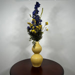 A yellow ribbed ceramic vase with a double-gourd shape, filled with a floral arrangement featuring dark blue delphiniums, yellow wildflowers, and green foliage, displayed on a dark brown wooden table against a plain light gray wall.