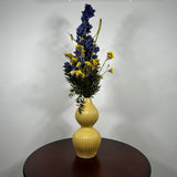 A yellow ribbed ceramic vase with a double-gourd shape, filled with a floral arrangement featuring dark blue delphiniums, yellow wildflowers, and green foliage, displayed on a dark brown wooden table against a plain light gray wall.
