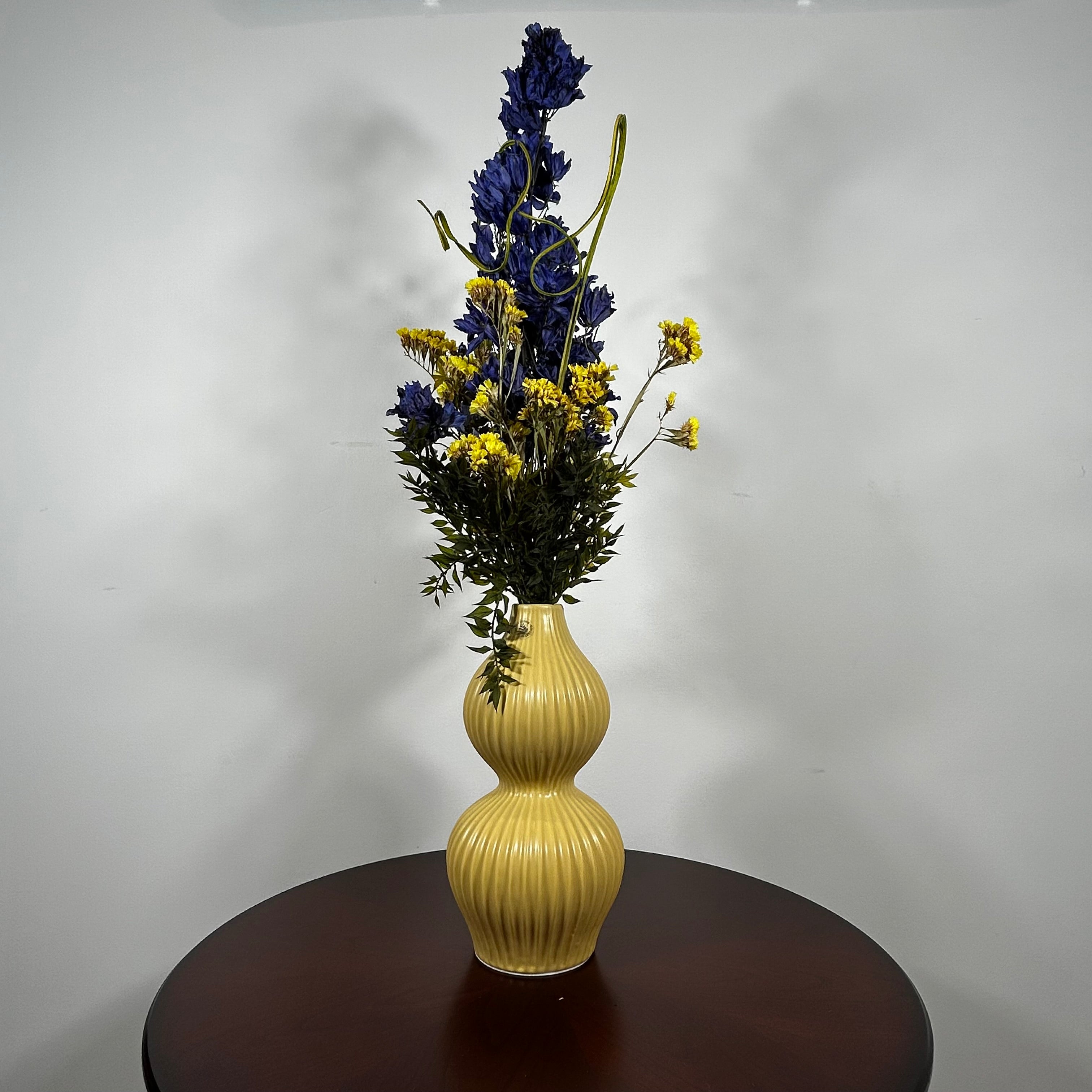 A yellow ribbed ceramic vase with a double-gourd shape, filled with a floral arrangement featuring dark blue delphiniums, yellow wildflowers, and green foliage, displayed on a dark brown wooden table against a plain light gray wall.