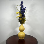 A two-tiered yellow ceramic vase with a ribbed texture, filled with a floral arrangement featuring blue delphiniums, yellow flowers, and green foliage, set on a dark wooden table against a plain white wall.