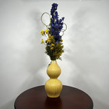 A two-tiered yellow ceramic vase with a ribbed texture, filled with a floral arrangement featuring blue delphiniums, yellow flowers, and green foliage, set on a dark wooden table against a plain white wall.