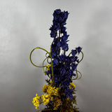A dried floral arrangement featuring tall dark blue delphiniums and clusters of small yellow flowers, accented with curly green stems, set against a plain light gray background.
