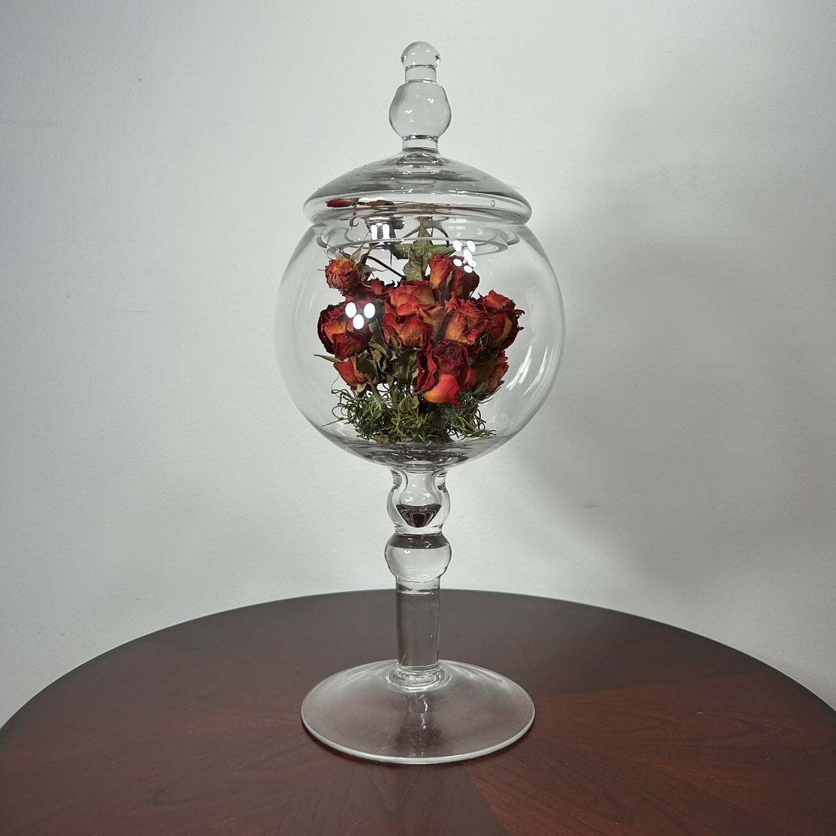 A clear glass apothecary-style jar with a lid and pedestal base, displayed on a dark wooden table against a light gray wall. Inside the glass sphere, dried red roses with green stems are arranged.