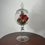 A clear glass apothecary-style jar with a lid and pedestal base, displayed on a round wooden table against a plain light gray wall. Inside the glass dome are dried red roses with green foliage.