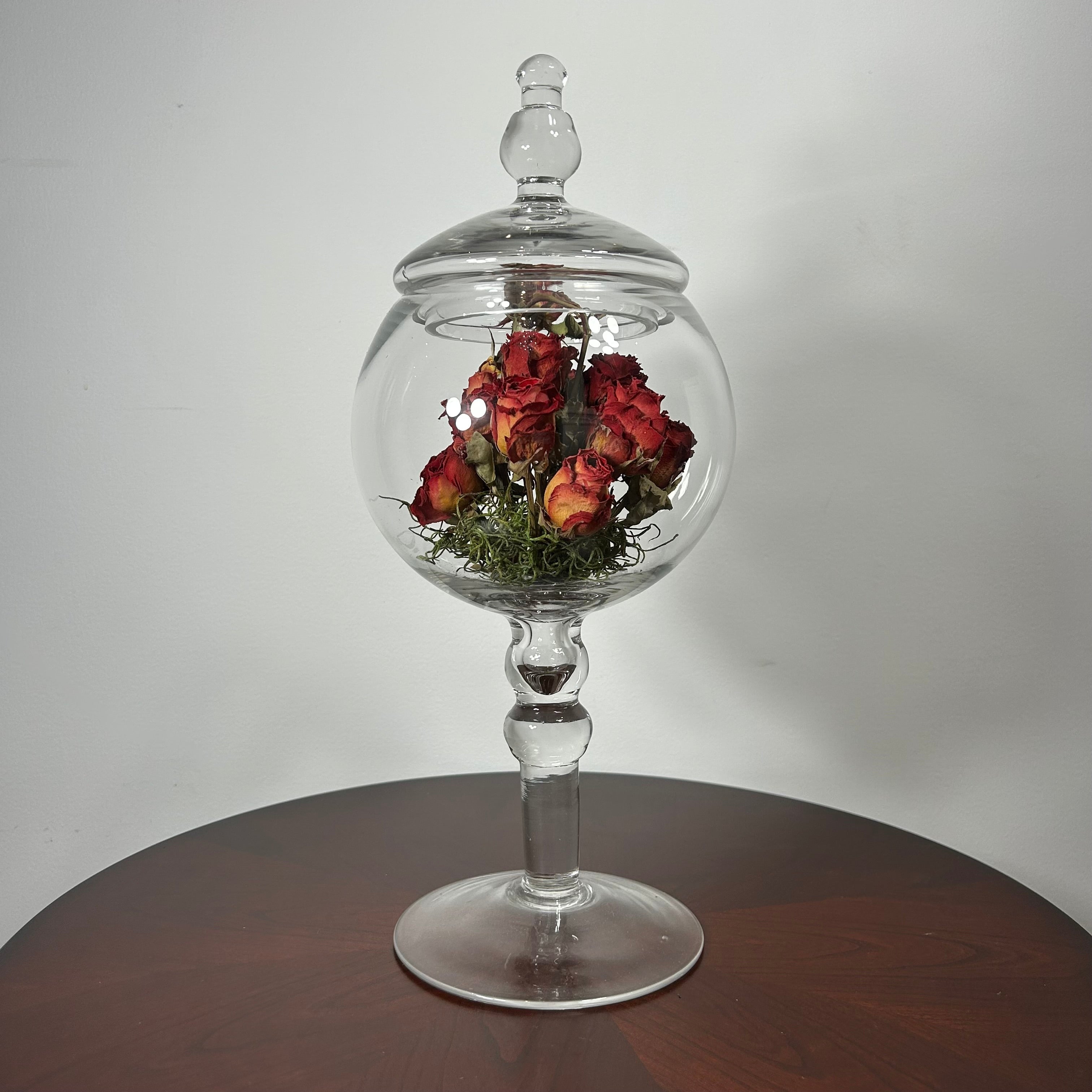 A clear glass apothecary-style jar with a lid and pedestal base, displayed on a round wooden table against a plain light gray wall. Inside the glass dome are dried red roses with green foliage.
