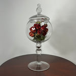 A clear glass apothecary-style jar with a lid and pedestal base, displayed on a round wooden table against a light gray wall. Inside the glass dome, dried red roses with green stems are arranged.