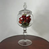 A clear glass apothecary-style jar with a lid and pedestal base, displayed on a round wooden table against a light gray wall. Inside the glass dome, dried red roses with green stems are arranged.